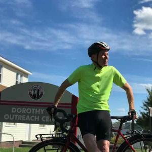 Brother Shamus McGrenra posing with his bike in front of Dorothy Day Outreach Center sign