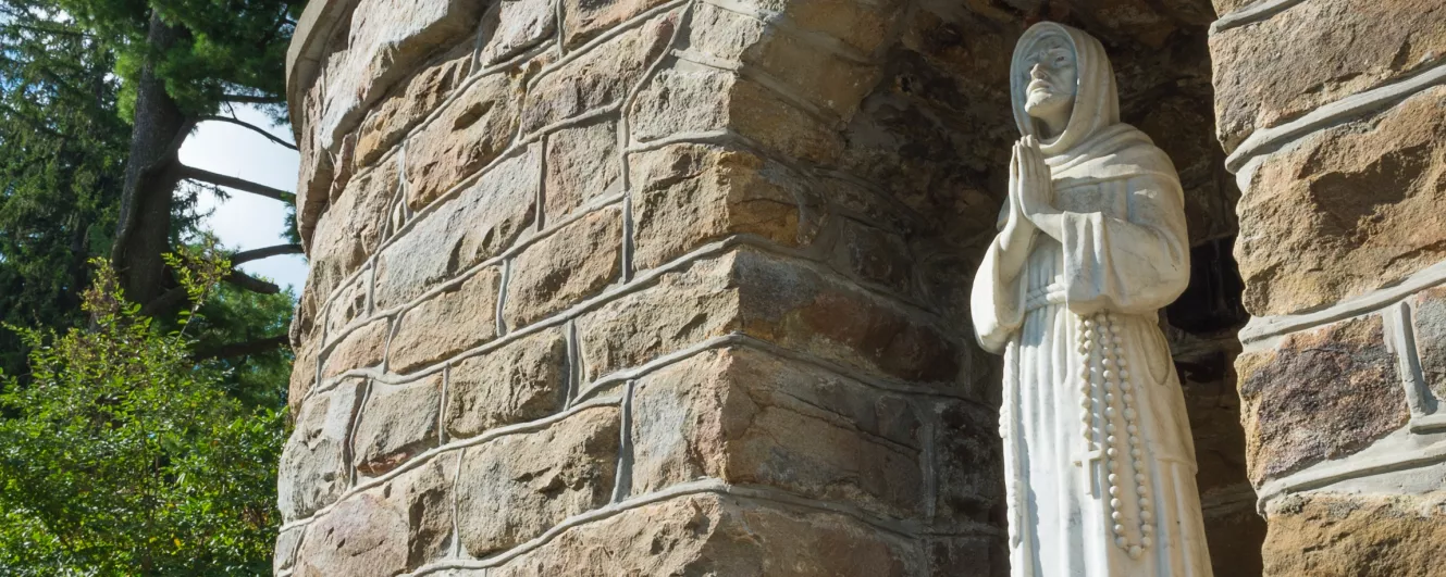 An Ancient stone wall hosting a hooded and pious Saint Francis gazing towards heaven. Sparsely garnished conifers glow green in the sunlight.